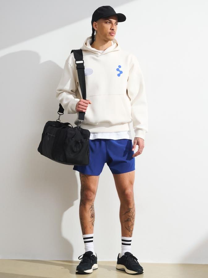 Person wearing a white hoodie with blue designs, blue shorts, and black sneakers, holding a black bag.
